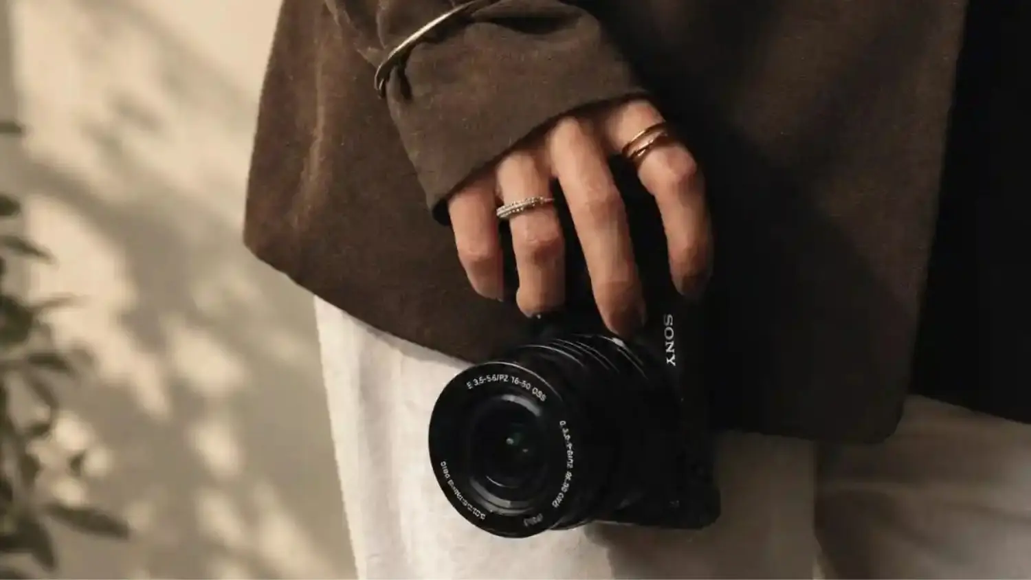 Person wearing a brown blazer and cream trousers holding a Sony mirrorless camera by their side.